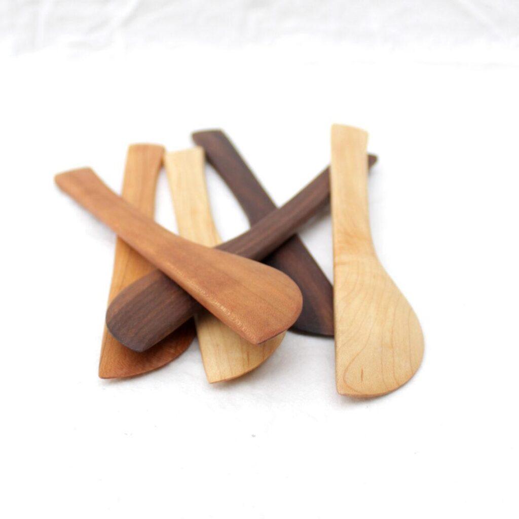 Five wooden butter knifes for crafting or cheese boards lie in a pile on top of a white cloth. They are made of salvaged/reclaimed hardwood: walnut, cherry, oak and maple. The picture is taken from the ends of the knives, so you can see how each one narrows to a pointy tip.