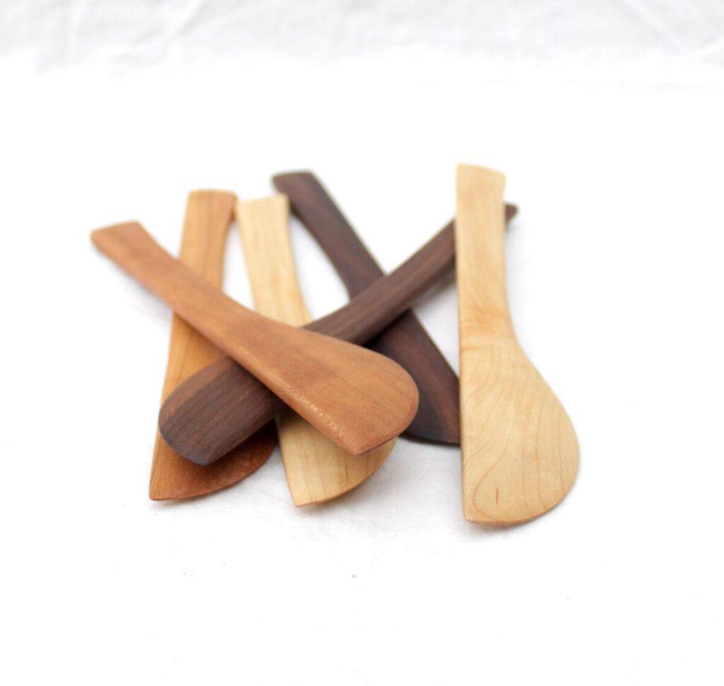Five wooden butter knifes for crafting or cheese boards lie in a pile on top of a white cloth. They are made of salvaged/reclaimed hardwood: walnut, cherry, oak and maple. The picture is taken from the ends of the knives, so you can see how each one narrows to a pointy tip.