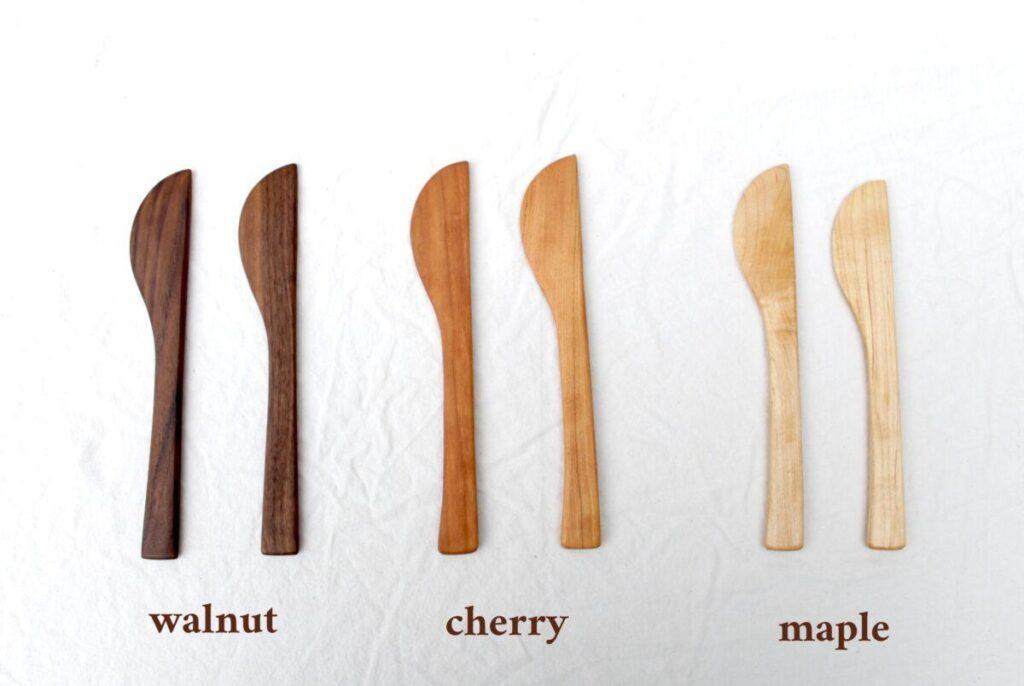 six pairs of cheese knives are lined up on white cloth. They are labeled with brown text below them to indicate the wood type. From left to right, the first pair is walnut, then cherry, then maple.