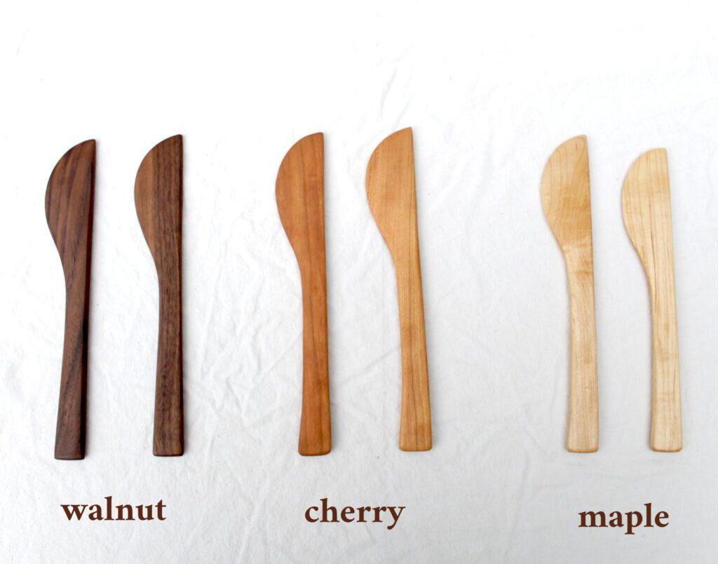 six pairs of cheese knives are lined up on white cloth. They are labeled with brown text below them to indicate the wood type. From left to right, the first pair is walnut, then cherry, then maple.