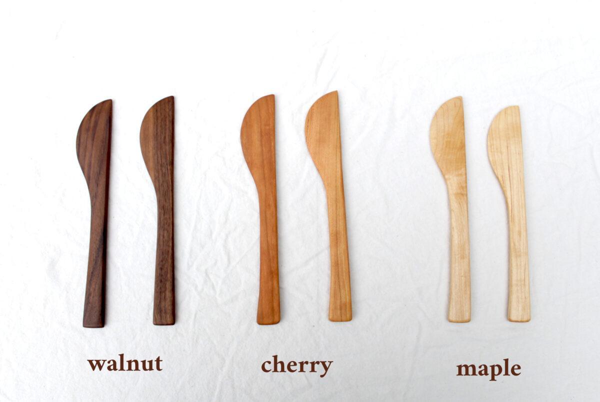 six pairs of cheese knives are lined up on white cloth. They are labeled with brown text below them to indicate the wood type. From left to right, the first pair is walnut, then cherry, then maple.
