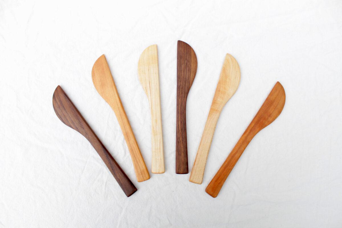 Top-down view of five butter, cheese or soap-beveling knives. They are arrayed in an arc, showing off the contrasting colors of the cherry, walnut and maply hardwoods. Each one is handmade and unique, but the overall shape is the same.
