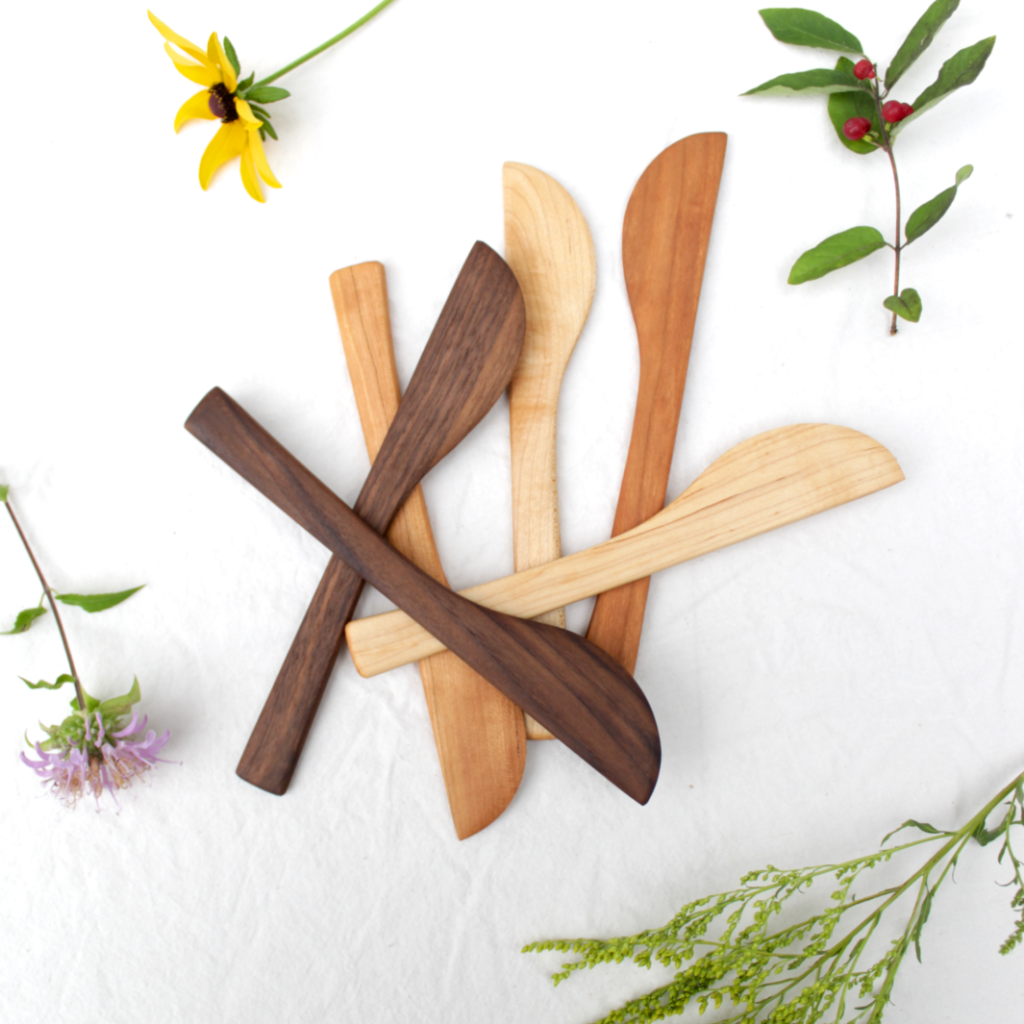 Six wooden butter knives are piled on a beige cloth with some flowers and plants strewn around them. The top two are walnut, then maple and cherry underneath. Each one is hand crafted by Flotsam.
