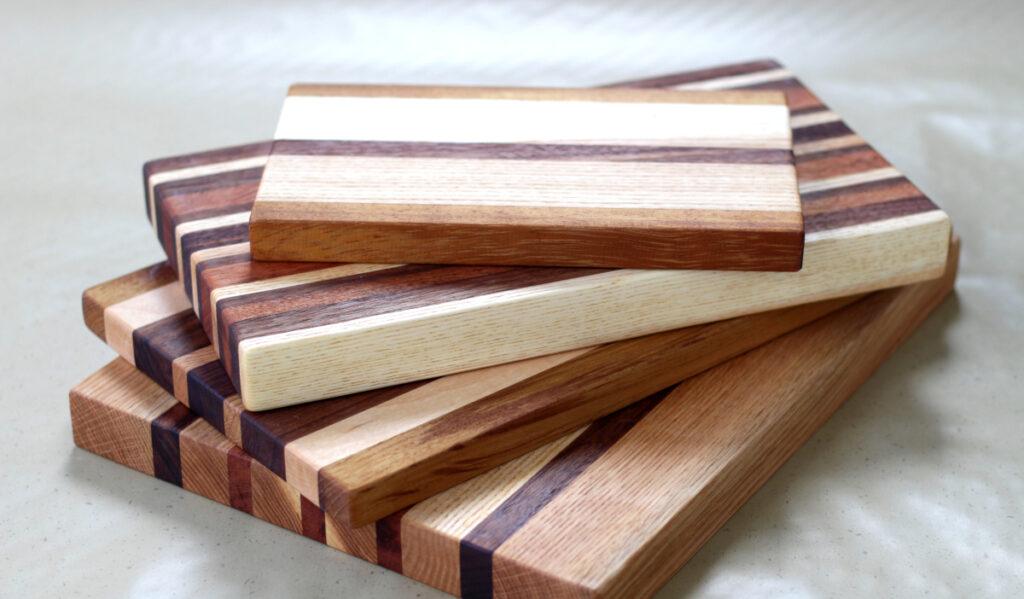 A stack of four custom-made limited-run cutting boards. They are all different sizes and thicknesses, and they are all made from strips of different sizes of contrasting wood. Handmade in the USA with salvaged materials.