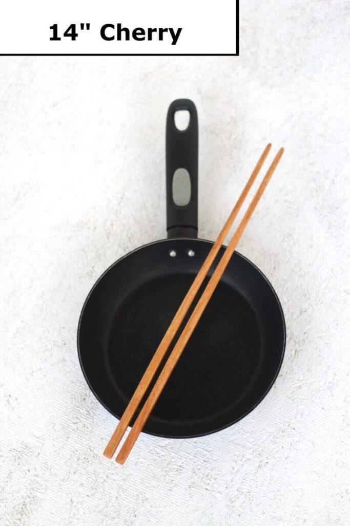 A pair of handmade wooden cooking chopsticks is lain across a small black frying pan. The label at the top reads "14 inch cherry". These kitchen utensils are made in the USA.