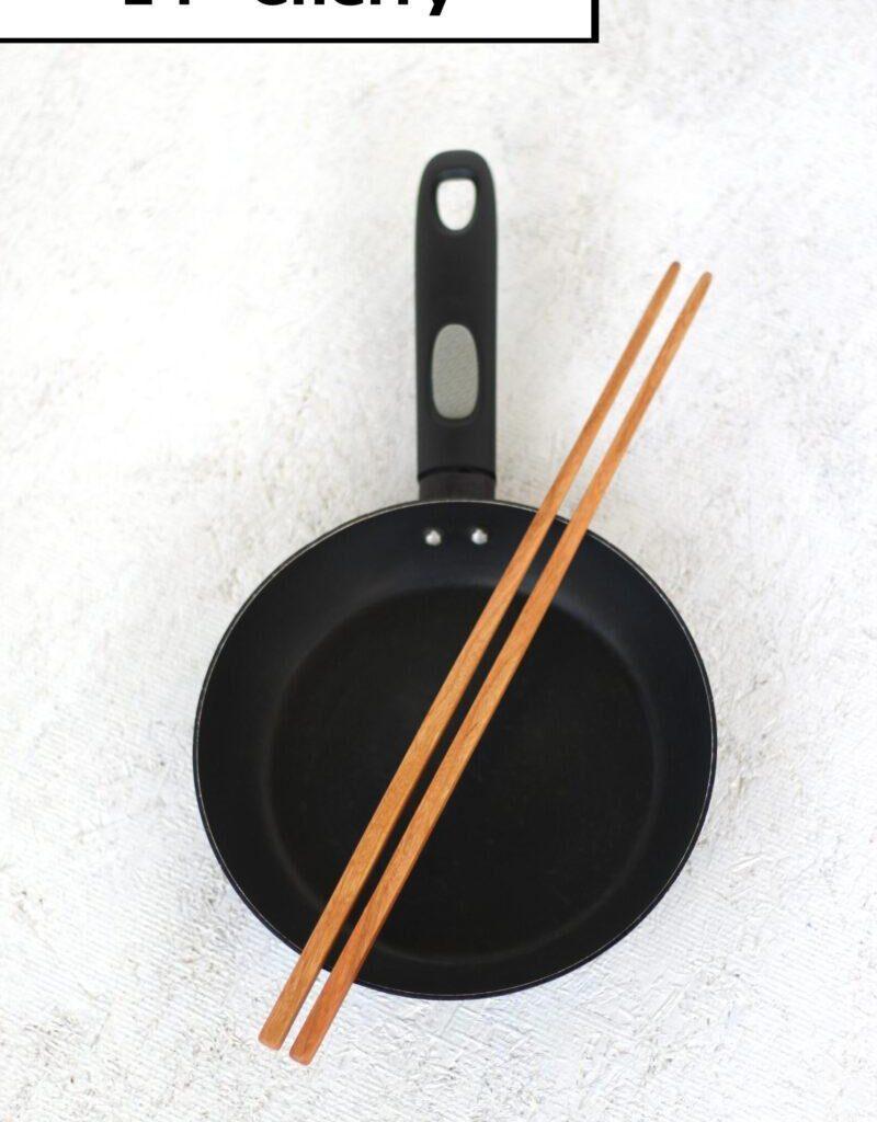 A pair of handmade wooden cooking chopsticks is lain across a small black frying pan. The label at the top reads "14 inch cherry". These kitchen utensils are made in the USA.