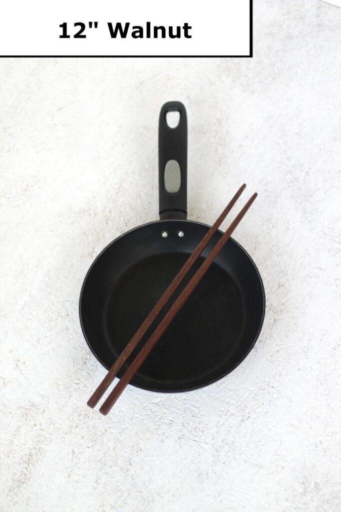 A pair of walnut chopsticks sits on top of a black frying pan, which sits atop a white speckled table. A lable at the top reads "12 inch Walnut".
