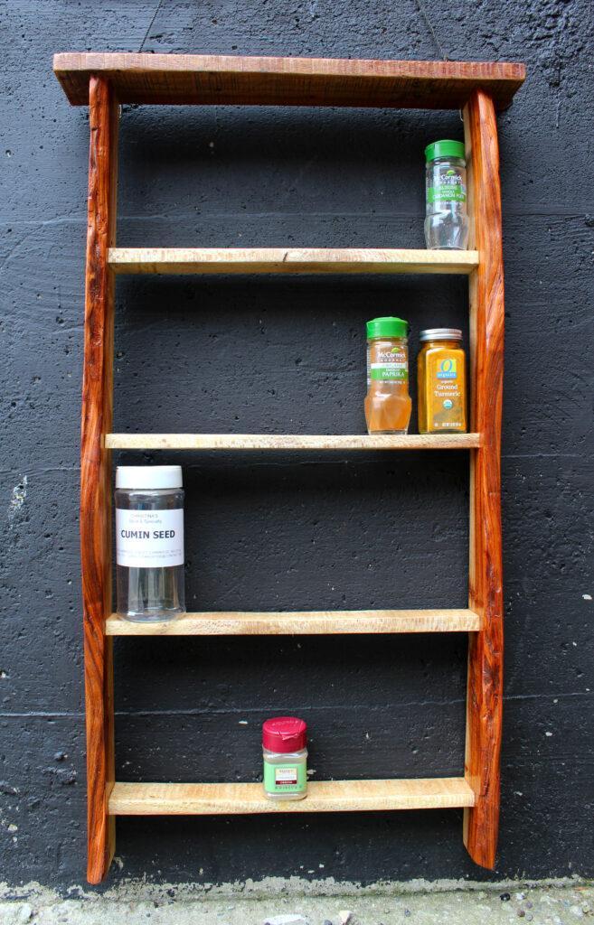 Hanging spice rack made from upcycled and reclaimed pallets. It has four shelves containing a few different herbs. Behind the shelf is a black cement wall. Made by Hudson Valley woodworker, Flotsam Made.