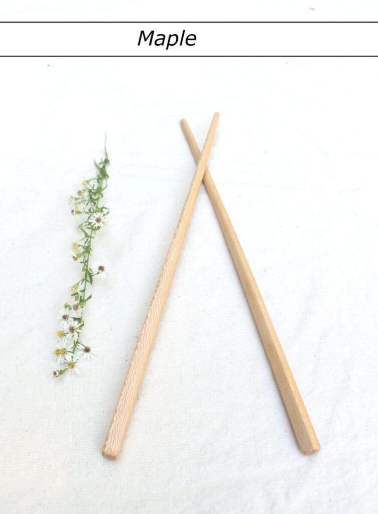 A single pair of light colored chopsticks lie with the tips crossed on a white tablecloth. A row of small white flowers is to the left. At the top is a label that reads "Maple". These hand crafted wooden utensils are made to last.