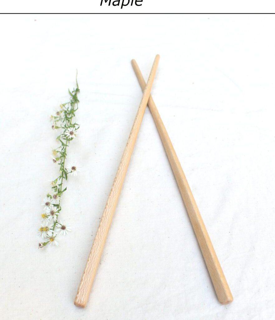 A single pair of light colored chopsticks lie with the tips crossed on a white tablecloth. A row of small white flowers is to the left. At the top is a label that reads "Maple". These hand crafted wooden utensils are made to last.