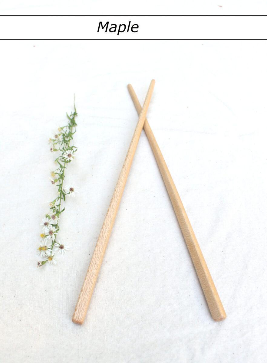 A single pair of light colored chopsticks lie with the tips crossed on a white tablecloth. A row of small white flowers is to the left. At the top is a label that reads "Maple". These hand crafted wooden utensils are made to last.