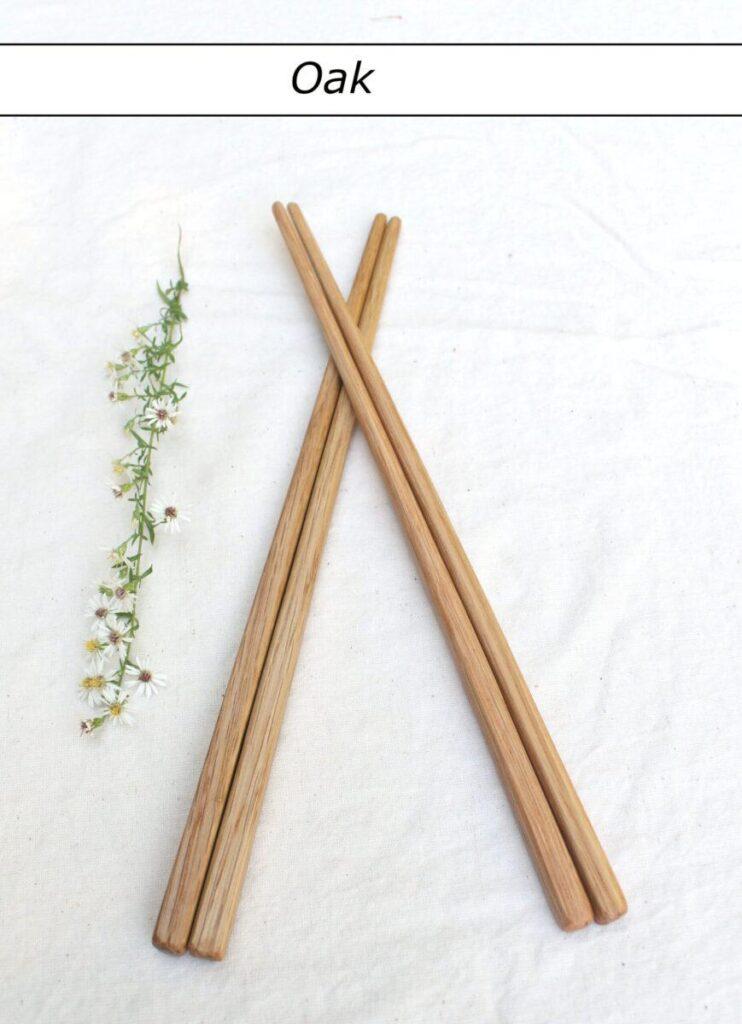 Two pairs of light-colored chopsticks lie on a white tablecloth with the tips crossed. To the left is a row of small white flowers. Above is a label that reads "Oak". These artisnal cooking tools have a durable and food-safe finish.