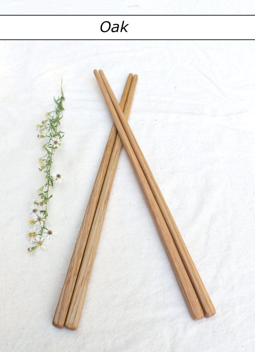 Two pairs of light-colored chopsticks lie on a white tablecloth with the tips crossed. To the left is a row of small white flowers. Above is a label that reads "Oak". These artisnal cooking tools have a durable and food-safe finish.