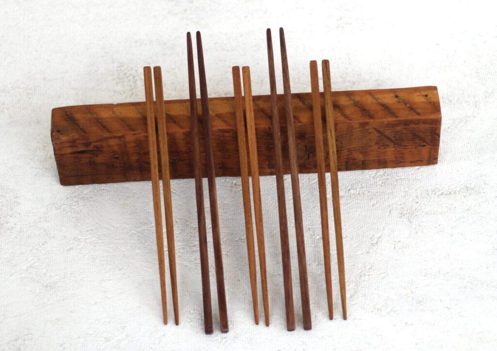 Five pairs of cooking chopsticks of different lenghts are lined up on a white table top, leaning on a short block of wood. The wood hue ranges from light reddish-brown to dark brown. Made with salvaged wood.