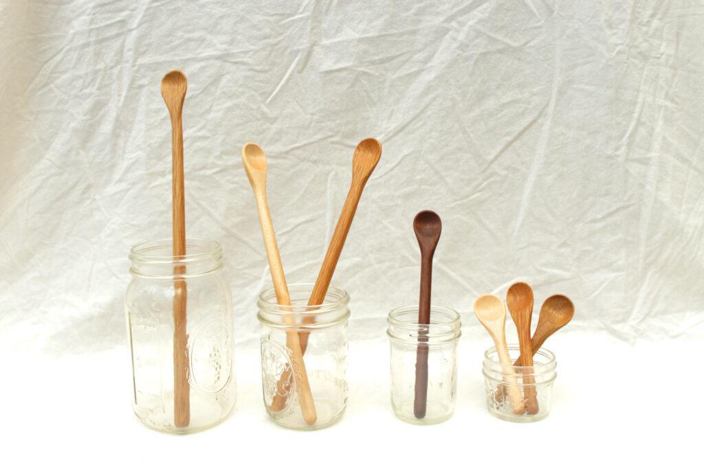 Four jars in a row hold different sized spoons. From left to right, the line of jars begins with a long mixing spoon in a quart jar. Then two shorter spoons in a pint jar, then one even shorter spoon in an 8 ounce jar, and finally three very short spoons in a 4 ounce jar.