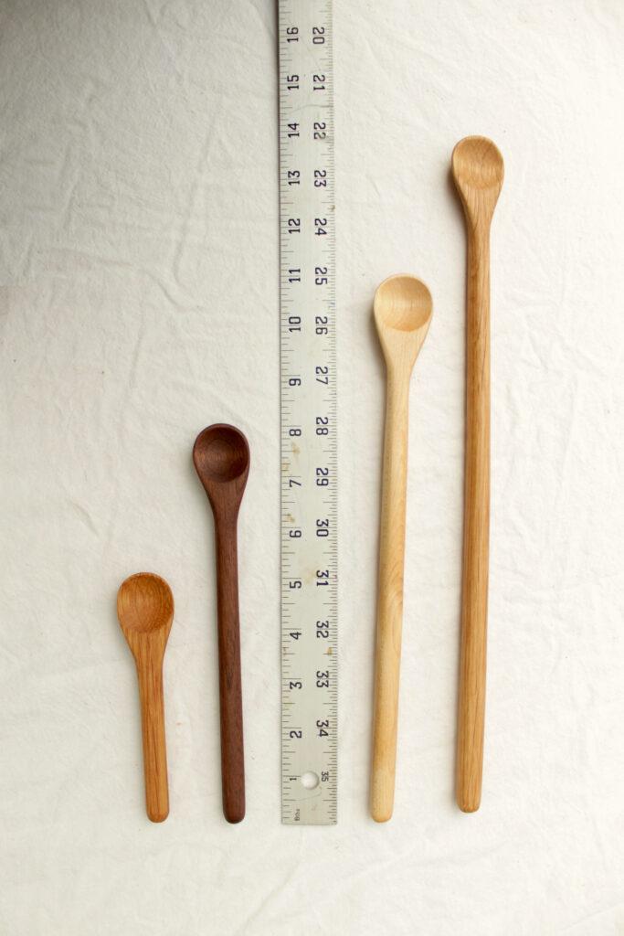 Four handmade stirring spoons lie on either side of a ruler. From left to right, the first stirring spoon is 5" cherry, then 8" walnut, then the ruler, then 11" maple, then 14" walnut. They are all hand crafted in the shawangunk mountains.