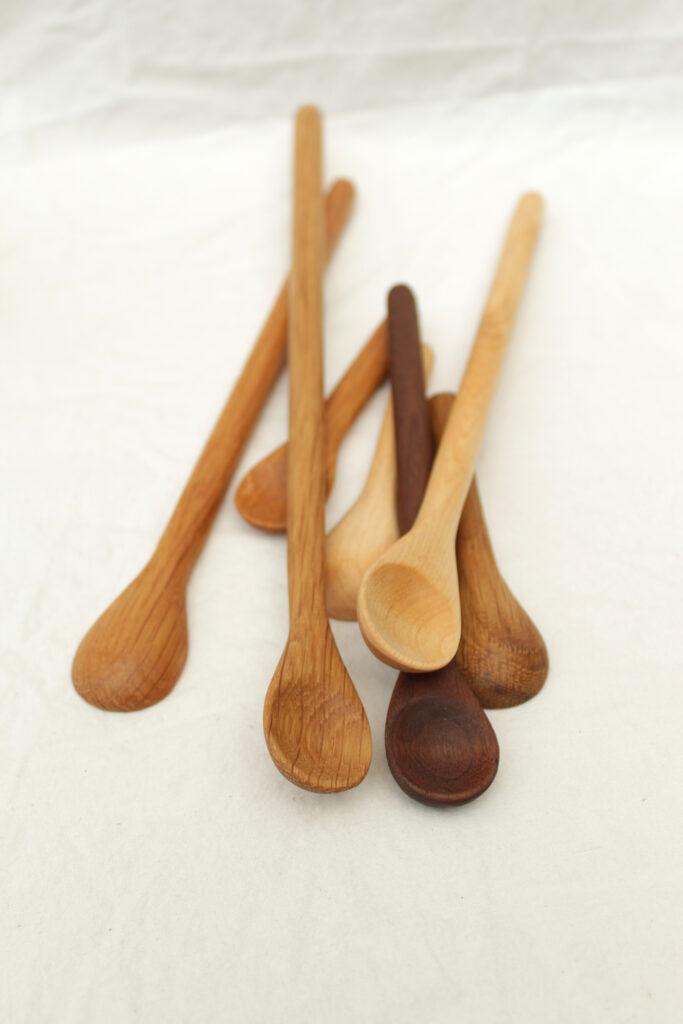 Seven artisnal wooden stirring spoons are lying in a messy stack on a cloth background. They are a mix of walnut, cherry, oak and maple and they are all different lengths. These are handmade in the catskills by a local maker.