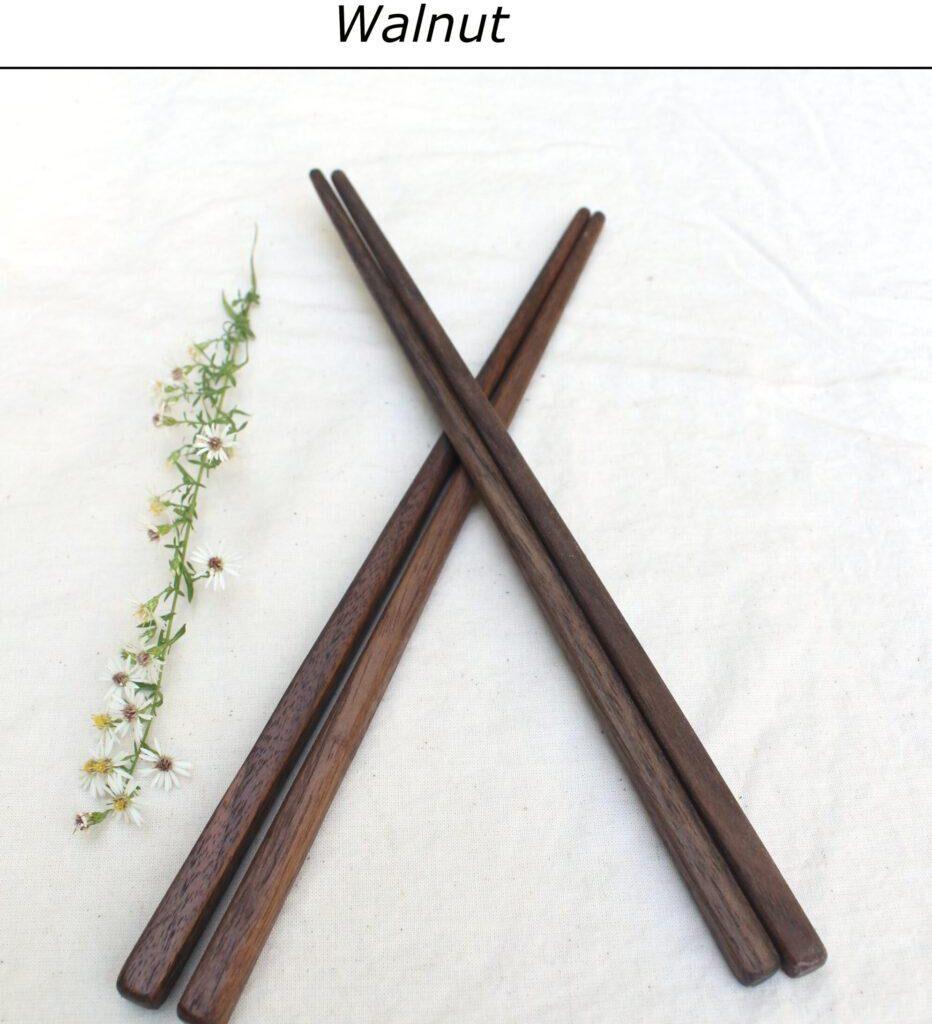 Two pairs of walnut chopsticks lie on a white table cloth with the tips crossing. A white flower is to the left of the chopsticks. A label with brown text says "Walnut" above them. Handmade in the USA.