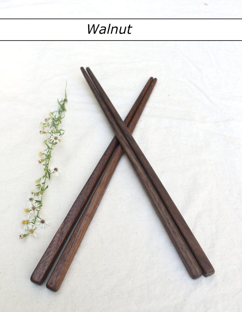Two pairs of walnut chopsticks lie on a white table cloth with the tips crossing. A white flower is to the left of the chopsticks. A label with brown text says "Walnut" above them. Handmade in the USA.