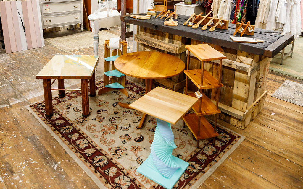An indoor booth displays a large table full of spoons, spatulas, butter knifes and cheese boards. In front of the display table is a large rug covering a hardwood floor, where a range of handmade one-of-a-kind side talbes and small shelves are arranged. Everything is handmade in the USA with salvaged wood.