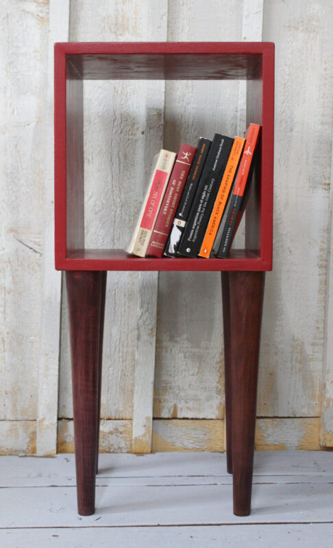 side table/bookshelf made from construction scraps A side table that doubles as a bookshelf. The top is an open red square with six books lined up inside. It sits on four dark-stained wood legs. This is handmade bespoke furniture by a Hudson Valley artisan.