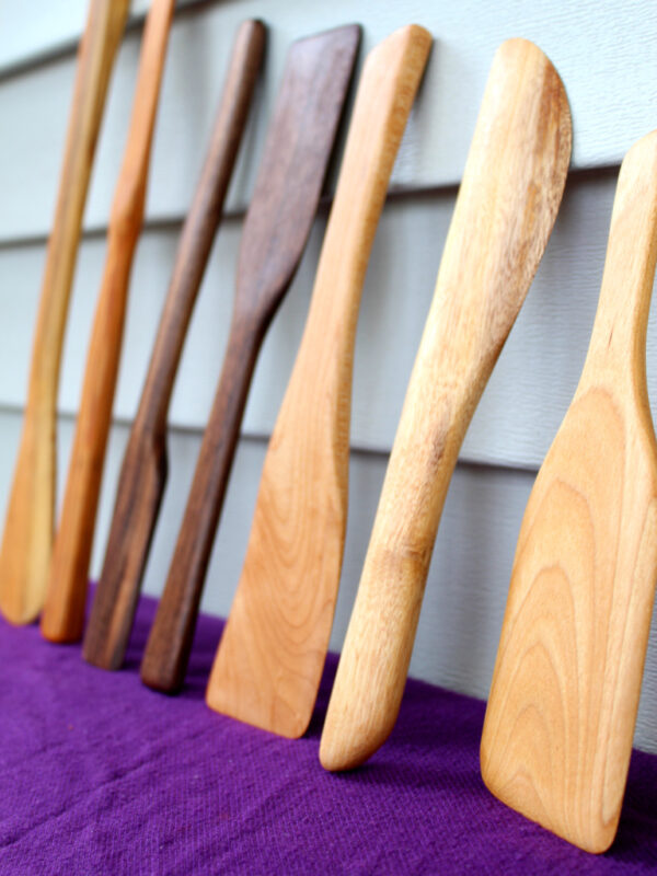 some of the earliest Flotsam spatulas, made from scraps (of course!) A line of hand-crafted small-batch spatulas made from upcycled and salvaged materials. There is a range of wood types, sizes and shapes. They are leaning vertically against a teal wall with a purple cloth underneath them. Each one is hand made in the USA.
